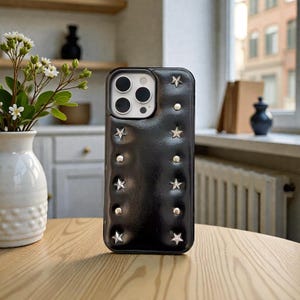 Puede incluir: Funda de teléfono negra con adornos de estrellas y tachuelas plateadas. La funda tiene una textura acolchada y un borde elevado alrededor de las lentes de la cámara. La funda está sobre una mesa de madera.