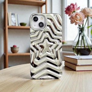 May include: Silver iPhone case with a star design. The case has a reflective, wavy pattern and a raised star in the center. The case is on a wooden table, with a bookshelf and flowers in the background.