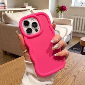 Puede incluir: Una funda de teléfono rosa brillante con un borde ondulado e irregular. La funda tiene un borde elevado alrededor de las lentes de la cámara. La funda del teléfono es de color sólido y se muestra en primer plano.