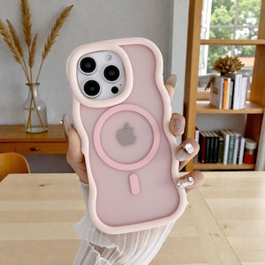 May include: A pale pink iPhone case with a wavy, light pink edge. The case features a circular MagSafe charger design and a camera cutout. The Apple logo is visible on the back of the case.