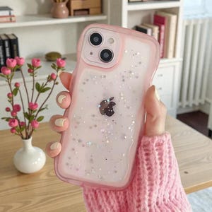 May include: A pink iPhone case with a clear back filled with glitter and star-shaped confetti. The case has a pink border and features the Apple logo. The phone case is held by a person wearing a pink jumper.
