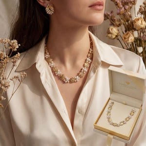 May include: Gold-toned necklace and matching earrings featuring a leaf design with small, round pearls. The necklace is displayed on a person wearing a white collared shirt. A jewelry box with the necklace inside is also visible.