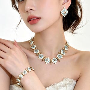 May include: A matching jewelry set featuring a necklace, bracelet, and earrings. The set has a gold-tone metal base with white enamel and blue floral accents. The earrings are a diamond shape. The necklace and bracelet have a repeating pattern.