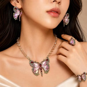 Könnte beinhalten: Ein Schmuckset mit Schmetterlingsmotiven, irisierenden rosa Flügeln, kleinen grünen und roten Kristallen sowie goldfarbenen Metallfassungen. Das Set besteht aus einer Halskette mit Perlenkette, passenden Ohrsteckern, einem Ring und einem Armband.