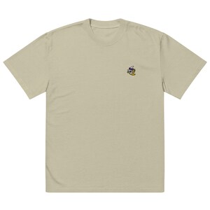Könnte beinhalten: Ein hellbeiges T-Shirt mit kurzen Ärmeln und einer kleinen gestickten Grafik auf der oberen linken Brustseite. Die Grafik zeigt eine gelbe Bananenfigur mit dunkler Sonnenbrille und weißem Hut in einer entspannten, geneigten Haltung.