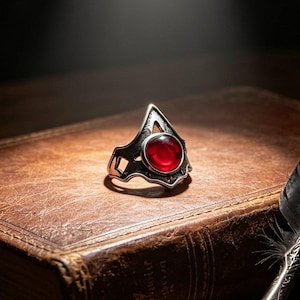 May include: A silver-toned ring featuring a prominent circular red gemstone set within an angular, openwork metal frame. The ring is positioned on a weathered, brown leather-bound book next to a black feather quill pen with a metallic nib.
