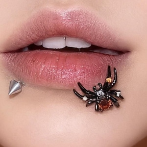 May include: Close-up of a person's lips with two pieces of jewelry. A silver spike stud is on the lower lip, and a black spider-shaped stud with red and clear gems is on the lower lip.