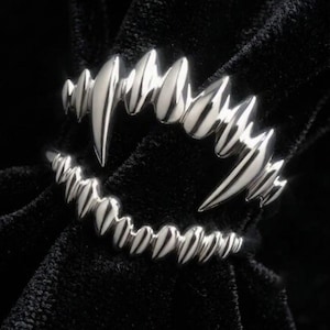 Puede incluir: Un anillo de tono plateado con un diseño de dos filas de formas puntiagudas similares a dientes. La fila superior tiene púas más grandes y alargadas, mientras que la fila inferior consta de puntos uniformes más pequeños en un aro abierto.