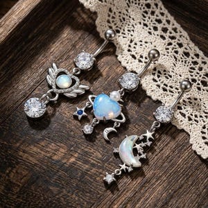 May include: Three silver-toned navel rings featuring celestial and heart motifs. The first has a heart with wings and a clear stone. The second features a heart-shaped opal-like stone with a small star. The third displays a crescent moon and stars.