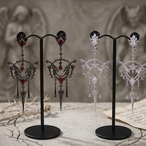 May include: Two pairs of ornate drop earrings displayed on black stands. One pair is black metal with deep red faceted stones, and the other is silver-toned metal with clear, light-reflecting stones. Both feature intricate wing and spike motifs.