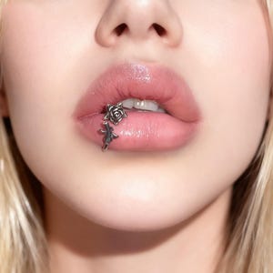 Könnte beinhalten: Nahaufnahme von Lippen mit einem silbernen Lippenpiercing. Das Piercing hat ein Rosen- und Stieldesign. Die Lippen sind glänzend und haben eine natürliche rosa Farbe.