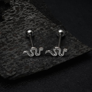 May include: Two silver-colored snake-shaped barbell earrings. Each earring features a small, round ball at the top and a detailed snake design at the bottom. The earrings are displayed on a dark, textured surface.