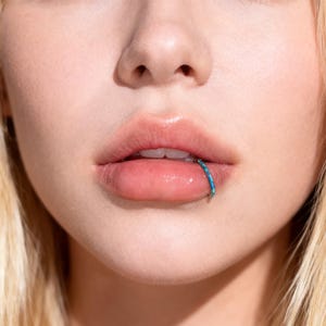 May include: Close-up of a person's lips with a blue and teal lip ring. The ring is a small hoop with a textured, iridescent appearance. The lips are full and glossy, with a natural pink hue.