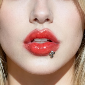 May include: Close-up of a person's lips with bright red lipstick and a small, silver cross stud piercing on the lower lip. The lips are glossy and the skin is fair.