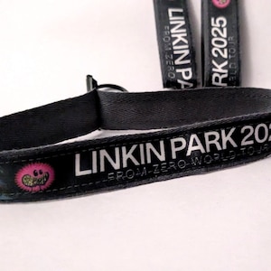 Könnte beinhalten: Schwarzes Stoffarmband mit weißem Text "LINKIN PARK 2025". Das Armband zeigt eine Cartoon-Grafik und die Worte "FROM-ZERO WORLD TOUR". Zwei passende schwarze Schlüsselbänder sind im Hintergrund zu sehen.