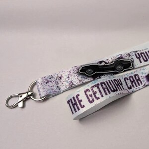 May include: Two white lanyards with silver-colored clasps. The lanyards have a purple and white splatter design and the text "YOU WERE DRIVING THE GETAWAY CAR" in purple. Each lanyard has a black car graphic.