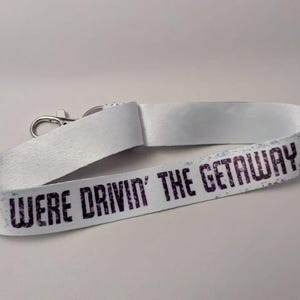 May include: White lanyard with the text "YOU WERE DRIVIN' THE GETAWAY CAR" in dark purple. The lanyard has a silver-colored metal clasp. The text has a distressed, vintage look.