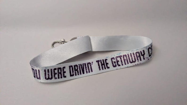 May include: White lanyard with the text "YOU WERE DRIVIN' THE GETAWAY CAR" in dark purple. The lanyard has a silver-colored metal clasp. The text has a distressed, vintage look.