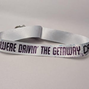 May include: White wristband with the text "YOU WERE DRIVIN' THE GETAWAY CAR" in dark purple. The wristband is made of a smooth, satin-like material and has a silver clasp.