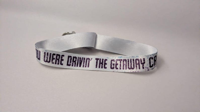 May include: White wristband with the text "YOU WERE DRIVIN' THE GETAWAY CAR" in dark purple. The wristband is made of a smooth, satin-like material and has a silver clasp.