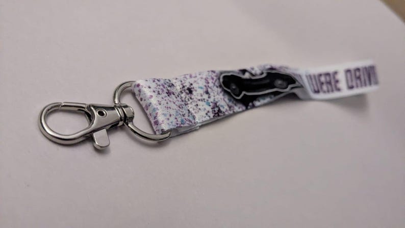 May include: A keychain with a silver clasp and a white strap. The strap has a black car graphic and a purple splatter design. The text "WERE DRIVIN" is printed on the end of the strap.