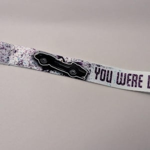 May include: A white lanyard with a black car graphic and the text "YOU WERE DRIVIN" in purple. The lanyard has a silver clasp and a speckled design in shades of purple and gray.