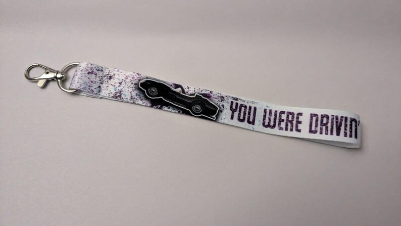 May include: A white lanyard with a black car graphic and the text "YOU WERE DRIVIN" in purple. The lanyard has a silver clasp and a speckled design in shades of purple and gray.
