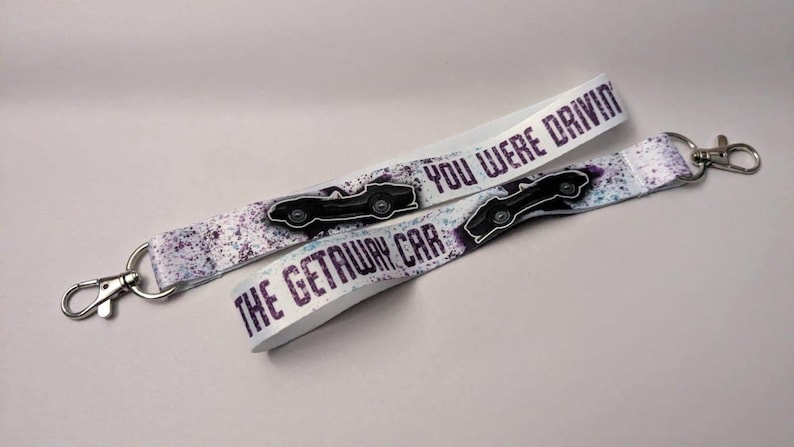 May include: Two white lanyards with silver-colored clasps. The lanyards have a purple and white splatter design and the text "YOU WERE DRIVING THE GETAWAY CAR" in purple. Each lanyard has a black car graphic.