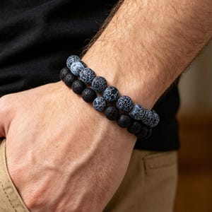 Könnte beinhalten: Ein zweireihiges Perlenarmband bestehend aus einer Reihe matter, schwarzer, poröser Lavasteinperlen und einer Reihe glatter, runder, blau-weißer Achatperlen mit Rissmuster, die auf einem flexiblen Gummiband aufgezogen sind.