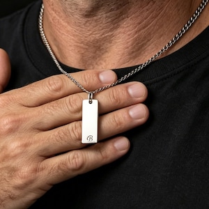 Könnte beinhalten: Halskette mit silberfarbenem, rechteckigem Anhänger und Kette. Der Anhänger hat einen kleinen, eingravierten Buchstaben "B" am unteren Rand. Die Halskette wird über einem schwarzen T-Shirt getragen.