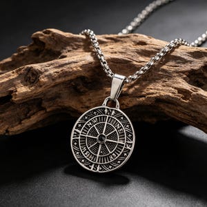 Pode incluir: Um colar com pingente prateado com um design de bússola circular. O pingente tem detalhes gravados intrincados e uma pequena pedra escura no centro. A corrente também é prateada e tem um design de elos. O fundo é um pedaço de madeira.