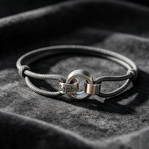 Könnte beinhalten: Ein graues Armband mit einem zentralen silberfarbenen Ring und einem Band mit dem Namen "Sophia". Das Armband hat ein geflochtenes Kordeldesign und ist vor einem dunkelgrauen Hintergrund platziert.