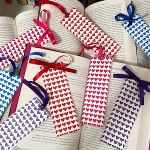 May include: Heart-patterned bookmarks in red, pink, blue, and purple, each with a matching ribbon. The bookmarks are placed among the pages of open books. A charming gift for book lovers.