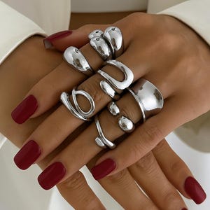Könnte beinhalten: Eine Sammlung silberner Ringe in verschiedenen abstrakten Designs. Die Ringe werden auf einer Hand mit rotem Nagellack gezeigt. Die Ringe haben ein glänzendes, metallisches Finish. Das Wort "ARTEVOA" befindet sich am unteren Rand des Bildes.