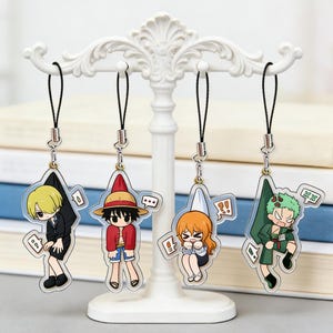 Może przedstawiać: Cztery breloczki z postaciami z anime wiszące na białym, dekoracyjnym stojaku. Breloczki przedstawiają Sanjiego, Luffy'ego, Nami i Zoro. Każda postać jest przedstawiona w stylu kreskówkowym z charakterystycznymi strojami i wyrazami twarzy. Breloczki są przymocowane do czarnych sznurków.
