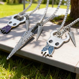 Puede incluir: Tres collares con colgantes plateados con diseños detallados. Uno presenta una cabeza blanca con ojos negros y una pluma azul y blanca, otro una daga plateada y el tercero una cabeza blanca con una pluma morada. Los collares se exhiben sobre una superficie de madera.