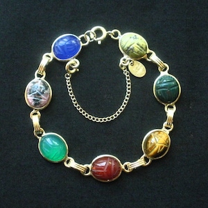 May include: A gold-tone link bracelet featuring seven oval-shaped scarab beetle charms in various colors including blue, green, red, yellow, and mottled patterns. The bracelet includes a small safety chain and a round metal tag with engraved text.
