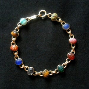 May include: A gold-toned chain bracelet featuring eleven oval-shaped, multi-colored gemstone scarab charms. Each charm is carved with a beetle design and linked together by small gold-toned metal rings, secured with a circular spring ring clasp.