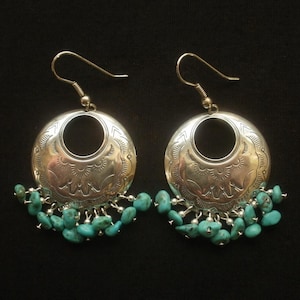 May include: A pair of silver-toned earrings with turquoise beads. Each earring features a circular design with an etched pattern and a dangling fringe of small, irregular turquoise beads. The earrings have a hook closure.