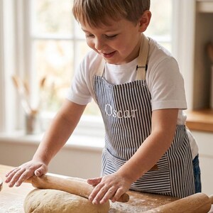 May include: A child wearing a striped apron with the name "Oscar" embroidered on it, rolling out dough on a wooden table. Cookie cutters in star and heart shapes are visible, along with a bowl of flour and rolling pins. The scene suggests baking.