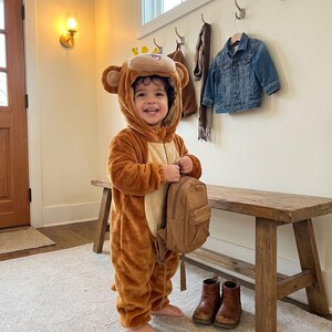 Puede incluir: Un ni&ntilde;o con un disfraz de mono marr&oacute;n con capucha, orejas y vientre marr&oacute;n claro. Sostiene una peque&ntilde;a mochila marr&oacute;n y lleva botas marrones. El fondo incluye un banco de madera, una chaqueta vaquera y una l&aacute;mpara de pared.