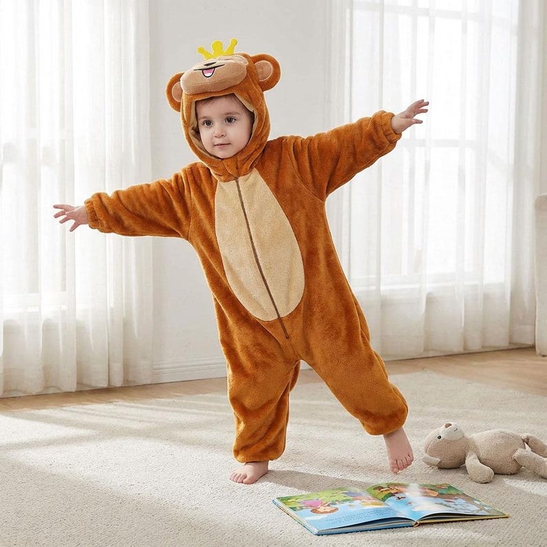 Puede incluir: Un ni&ntilde;o con un disfraz de mono marr&oacute;n con capucha y corona amarilla. El disfraz tiene una barriga marr&oacute;n claro y una cremallera. El ni&ntilde;o est&aacute; de pie sobre una alfombra clara con los brazos extendidos. Un peluche y un libro est&aacute;n en el suelo.