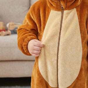 Puede incluir: Un mono marr&oacute;n de felpa con capucha y vientre de color beige claro. El mono tiene una cremallera en la parte delantera y est&aacute; hecho de un material suave y difuso. La mano de un ni&ntilde;o sostiene el lateral del mono.