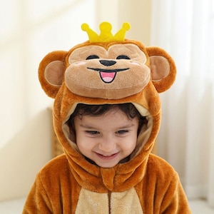 Puede incluir: Un ni&ntilde;o con un disfraz de mono marr&oacute;n con una corona amarilla en la capucha. El disfraz tiene una cara de mono sonriente con orejas grandes. El ni&ntilde;o sonr&iacute;e y mira hacia abajo.