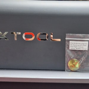 Puede incluir: Primer plano de una máquina gris oscuro con la palabra "XTOOL" en letras plateadas. Una pequeña bolsa de plástico contiene una lente de enfoque. La lente es circular y tiene una apariencia iridiscente dorada y verde. El texto en la bolsa dice "Tool P2/P25 Focusing Lens".