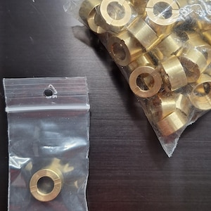 Puede incluir: Una colección de anillos metálicos de color latón, algunos en una bolsa de plástico transparente. Los anillos tienen forma circular con una ranura en un lado. Los anillos probablemente se utilizan para fines mecánicos o industriales.