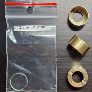 Puede incluir: Tres casquillos de latón y una bolsa de plástico transparente con un cierre rojo. La bolsa contiene un objeto pequeño, plano y circular. La etiqueta de la bolsa dice "ø12,5 mm x 1 mm". Los casquillos son cilíndricos con un agujero central.