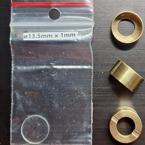 Puede incluir: Una bolsa de plástico transparente con cremallera roja contiene un objeto pequeño, transparente y circular. La bolsa tiene el texto "ø13,5mm x 1mm". También se ven tres anillos y cilindros metálicos de color latón.