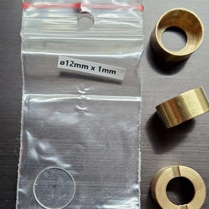 Pode incluir: Um saco plástico transparente contendo um pequeno disco transparente e uma etiqueta com o texto "ø12mm x 1mm". Três anéis cilíndricos de latão também são visíveis, com um anel tendo uma ranhura. Os itens estão em uma superfície escura.