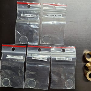 Puede incluir: Cinco bolsas de plástico transparente con cierres rojos, cada una con un objeto circular pequeño y transparente. Las bolsas tienen etiquetas que dicen "ø13mm x 1mm". También se ven varios objetos cilíndricos de color latón.